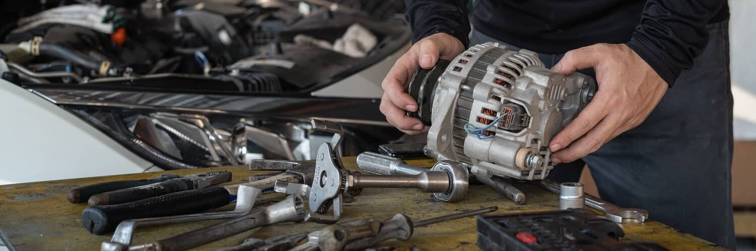 Technician holding a removed alternator beside workshop tools