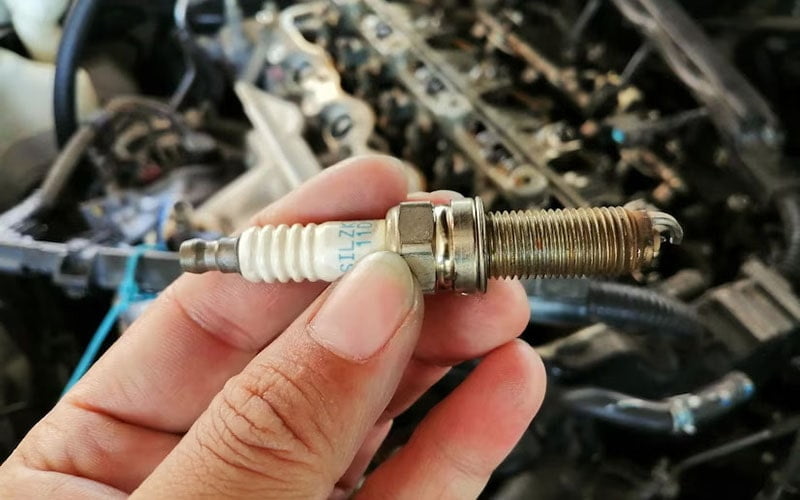 A spark plug held beside an open engine bay