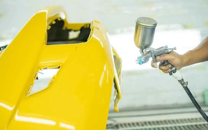 A spray gun applying yellow paint to a removed bumper cover